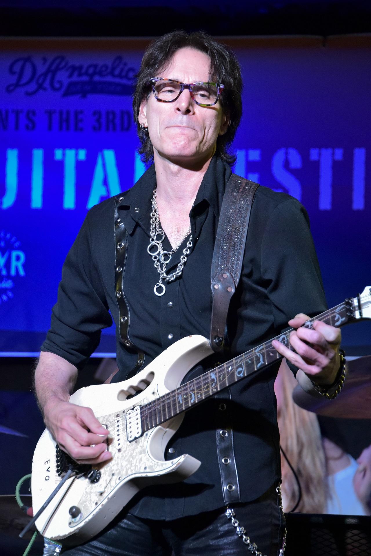 A man wearing glasses plays guitar against a blue background.